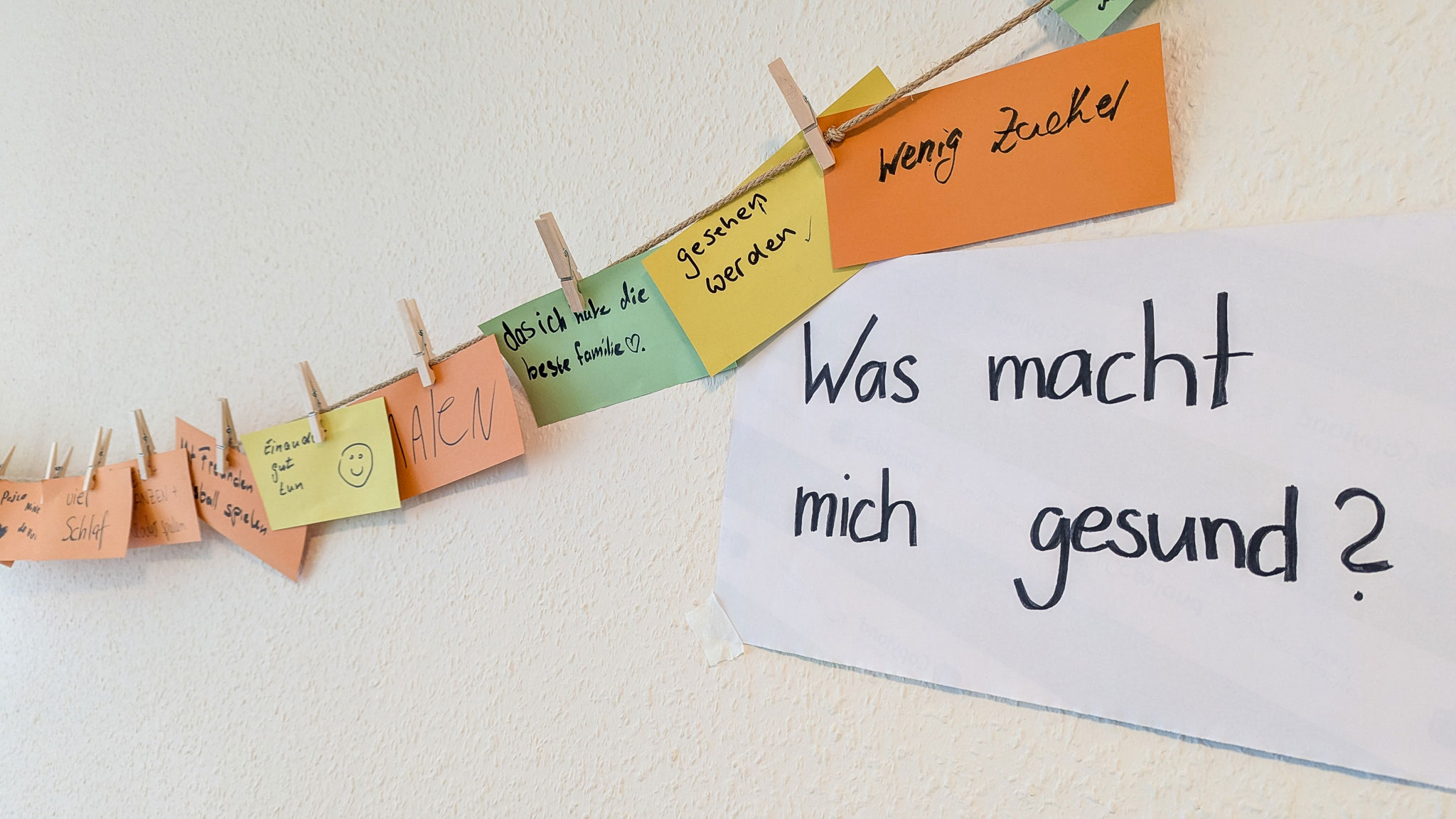 Vor einer Wand hängt eine Leine, an der Zettel hängen, auf die Menschen geschrieben haben, was sie gesund macht. Zu lesen ist “wenig Zucker”, “gesehen werden”, “das ich habe die beste Familie”, “Malen”, “einander gut tun”, “mit Freunden Fußball spielen” und “viel Schlaf”